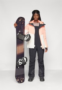 Snowboard con un top viola scuro e attacchi bianchi, tenuto da una persona in giacca multicolore, tute nere e occhiali da sci.