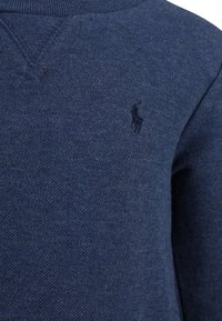 Mörkblå stickad tröja med en texturerad yta, som har en liten broderad logotyp på bröstet och en v-formad halsringning.