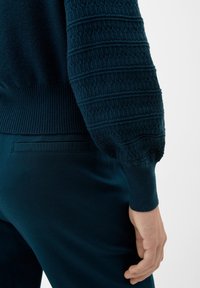 Ciemnozielony sweter dziany z teksturowanymi puff sleeves i ściągaczami, w połączeniu z ciemnozielonymi spodniami z tylną kieszenią. Widoczna miękka tkanina.