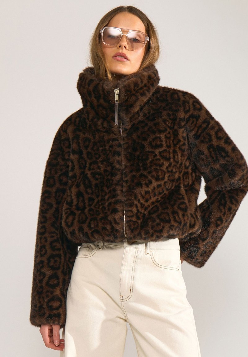 Femme portant une veste oversize en fausse fourrure marron à imprimé léopard avec col haut et pantalon beige clair, posant avec une main sur la hanche.