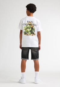 T-shirt blanc en coton avec un motif coloré de cactus dans le dos, associé à un short en denim foncé et des baskets blanches.