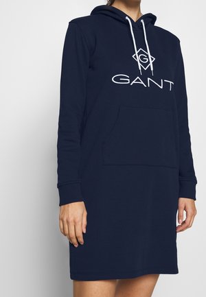 Woman wearing navy blue hoodie dress with front pocket and white GANT logo and drawstrings, shown from neck to mid-thigh.