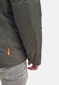 Giubbotto imbottito verde oliva con texture liscia, dotato di un'etichetta logo arancione visibile sul lato sinistro. Jeans grigi visibili di sotto.