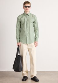 Jovem vestido com uma camisa de botão verde clara, calças largas bege, sapatos pretos, óculos de sol escuros, segurando uma grande bolsa tote preta.