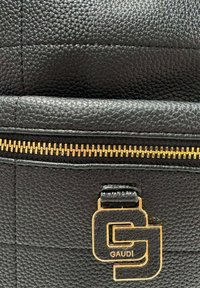 Dettaglio di una borsa in pelle nera testurizzata con cerniera dorata e logo metallico a forma di "G" sovrapposto con la scritta "GAUDI" sul fronte.