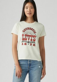 Camiseta de algodón color beige claro con un estampado gráfico vintage rojo que dice "AUTHENTIC LEVI'S", con cuello redondo y mangas cortas.