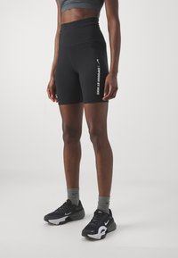 Czarne wysokie spodenki rowerowe o gładkiej fakturze z białym logo "Swoosh". Noszone z czarnymi butami sportowymi i szarymi skarpetkami.