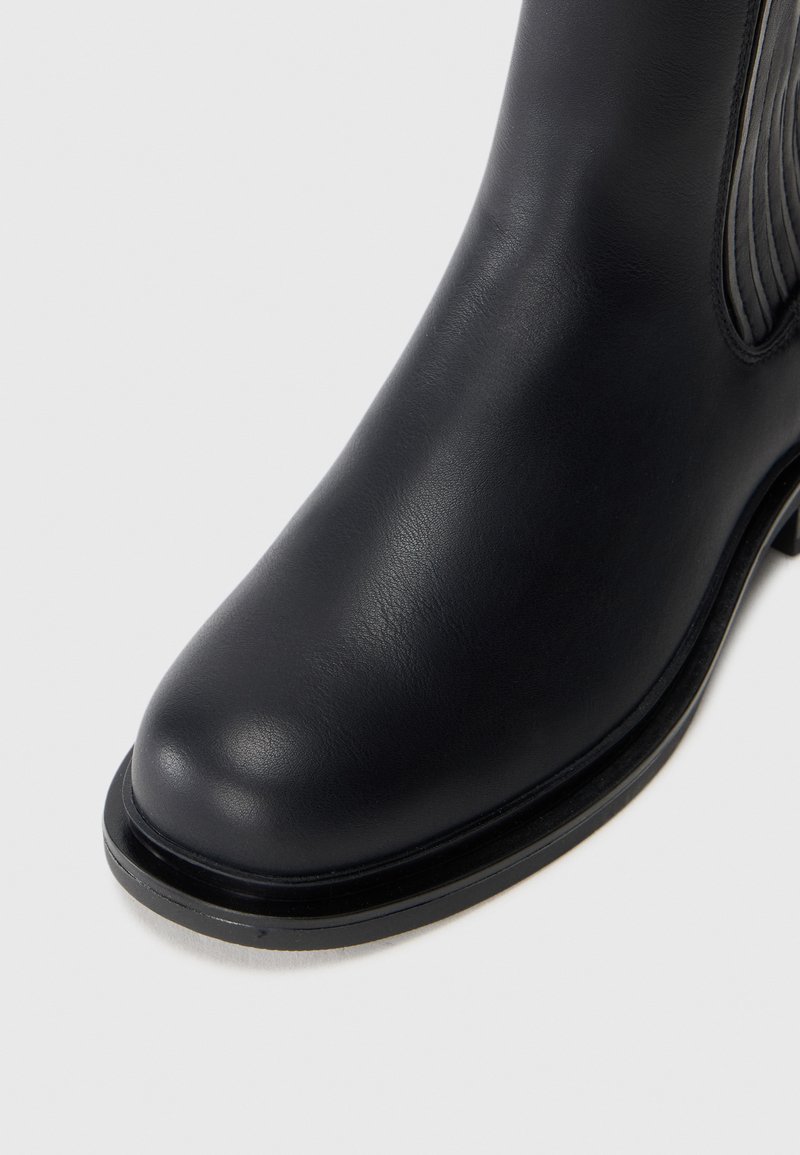 Stivale alla caviglia in pelle nera con finitura liscia, punta arrotondata e una sottile cucitura decorativa lungo il lato. Suola robusta e design minimalista.