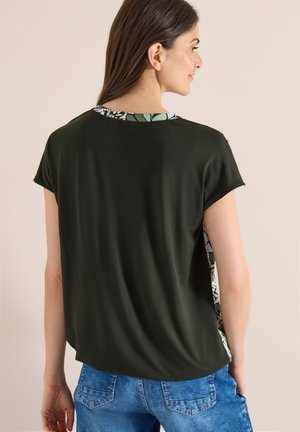 Femme vue de dos portant un haut noir à manches courtes avec bordure florale et un jean en denim bleu sur un fond uni.