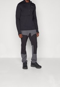 Zwarte pullover met lange mouwen en een rits bij de hals, gecombineerd met grijs en zwart gepaneerde broeken en zwarte sportschoenen. Glad stoftextuur.