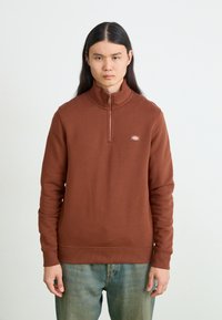 Smeđa zip-up dukserica s visokim ovratnikom, sa rebrastim manžetama i donjim rubom, koja sadrži mali logo na prsima. Uparena s plavim trapericama.
