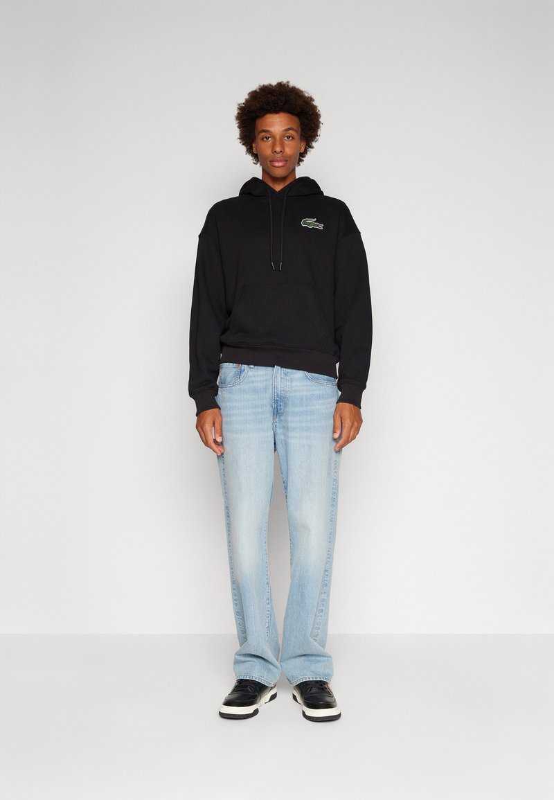 Felpa nera in cotone con tasca frontale, caratterizzata da un piccolo logo verde. Jeans in denim azzurro chiaro, leggermente svasati in fondo. Scarpe nere.
