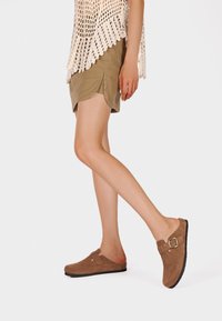 Scarpe slip-on in suede marrone con punta arrotondata, con un dettaglio a fibbia laterale e una superficie testurizzata. Indossate con una gonna kaki.