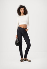 Μαύρα skinny τζην με λεία υφή denim, συνδυασμένα με ένα λευκό κοντό μακρυμάνικο μπλουζάκι και παπούτσια με λεοπάρ σχέδιο, κρατώντας μία μικρή μαύρη τσάντα.