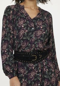 Schwarzes Blumenkleid mit langen Ärmeln, das lila und grüne Rosenmuster aufweist, einen Kragen hat und einen breiten schwarzen Gürtel mit goldenen Nieten trägt.