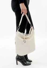Sac seau en cuir blanc avec une fermeture à cordon, quincaillerie en argent et une bandoulière amovible, tenu par une personne portant un jean noir et des bottes.