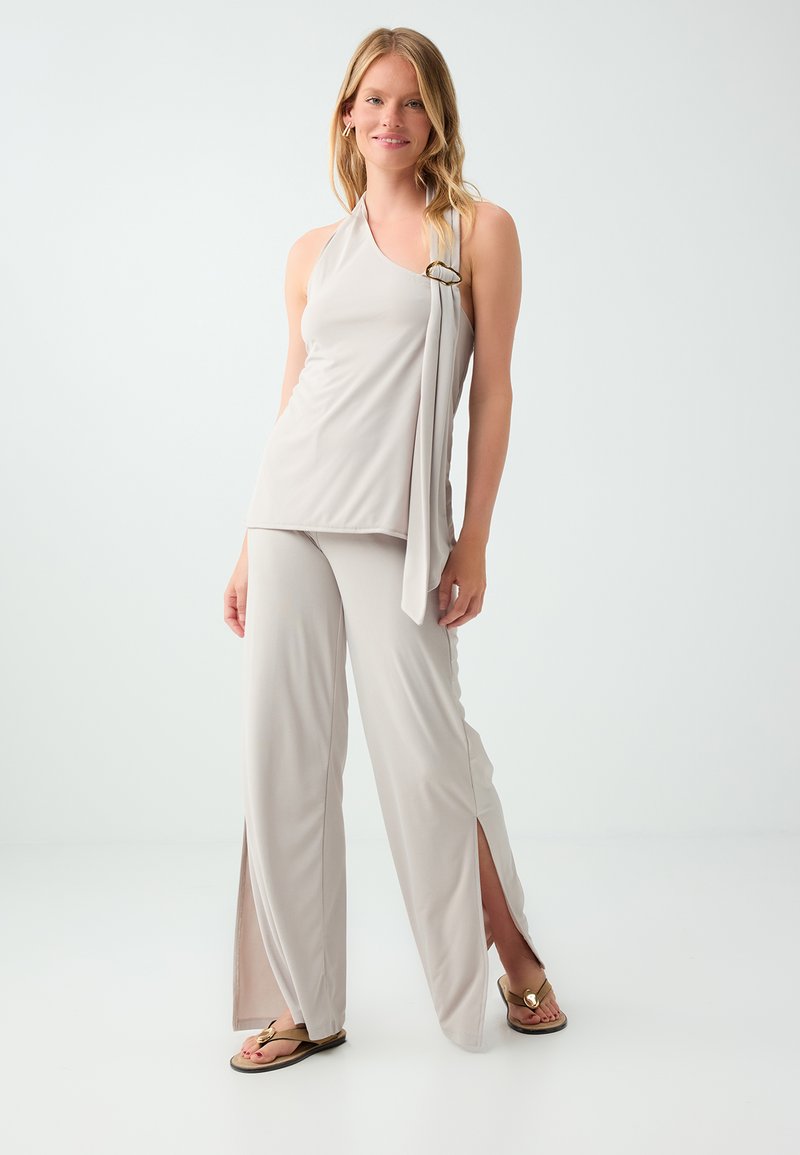 Femme portant un haut halter sans manches beige clair avec une ceinture et un pantalon à jambes larges avec des fentes à l’avant, debout sur un fond blanc uni.
