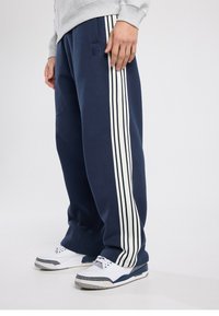 Pantalon de jogging bleu marine avec des bandes blanches sur les côtés, taille côtelée, deux poches latérales et un logo sur la cuisse gauche.