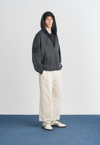 Γκρι φούτερ με φερμουάρ και μπροστινή τσέπη, συνδυασμένο με λευκά φαρδιά παντελόνια και λευκά αθλητικά παπούτσια με μαύρες ρίγες.