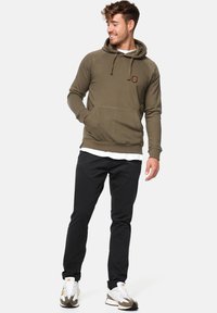 Sudadera verde oliva con un bolsillo frontal y capucha ajustable, combinada con pantalones negros y zapatillas deportivas blancas.