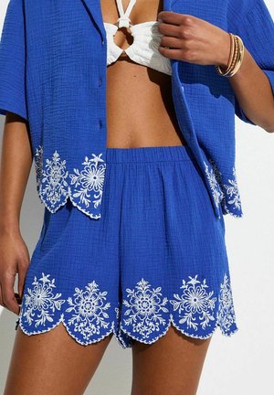 Femme portant un short bleu texturé et une chemise à manches courtes assortie avec une broderie florale blanche, associée à un haut de bikini blanc à col licou.
