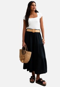 Gonna lunga nera a balze con una cintura intrecciata beige, abbinata a una maglietta bianca con scollatura quadrata e sandali neri con tacco alto. Gli accessori includono una borsa a cestino intrecciata.