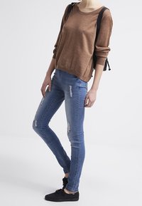 Jersey marrón de manga larga con un corte relajado. Jeans skinny desgastados de un azul claro. Zapatillas negras. La modelo lleva una mochila negra.