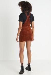 Robe salopette en velours côtelé de couleur rouille, avec une coupe ajustée, des bretelles réglables, une poche arrière et un ourlet effiloché. Associée à des bottes noires.