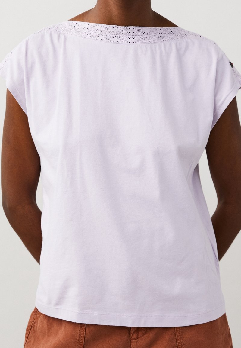 Osoba oblečená v světle fialovém tričku s krátkým rukávem a krajkovým detailem na ramenou a hnědých kalhotách, vyfotografována zezadu.