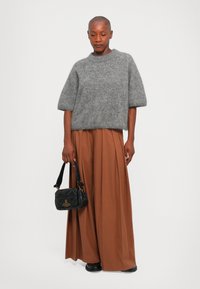 Personne portant un pull gris à manches courtes, un pantalon large plissé marron, des chaussures noires, et tenant un petit sac à main noir sur fond uni.