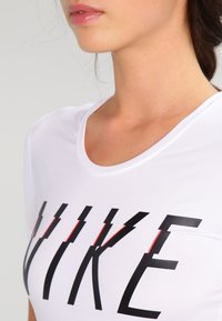 Camiseta Nike blanca con cuello redondo, que presenta letras negras en negrita y acentos rojos en la "K", hecha de un material suave y liviano.