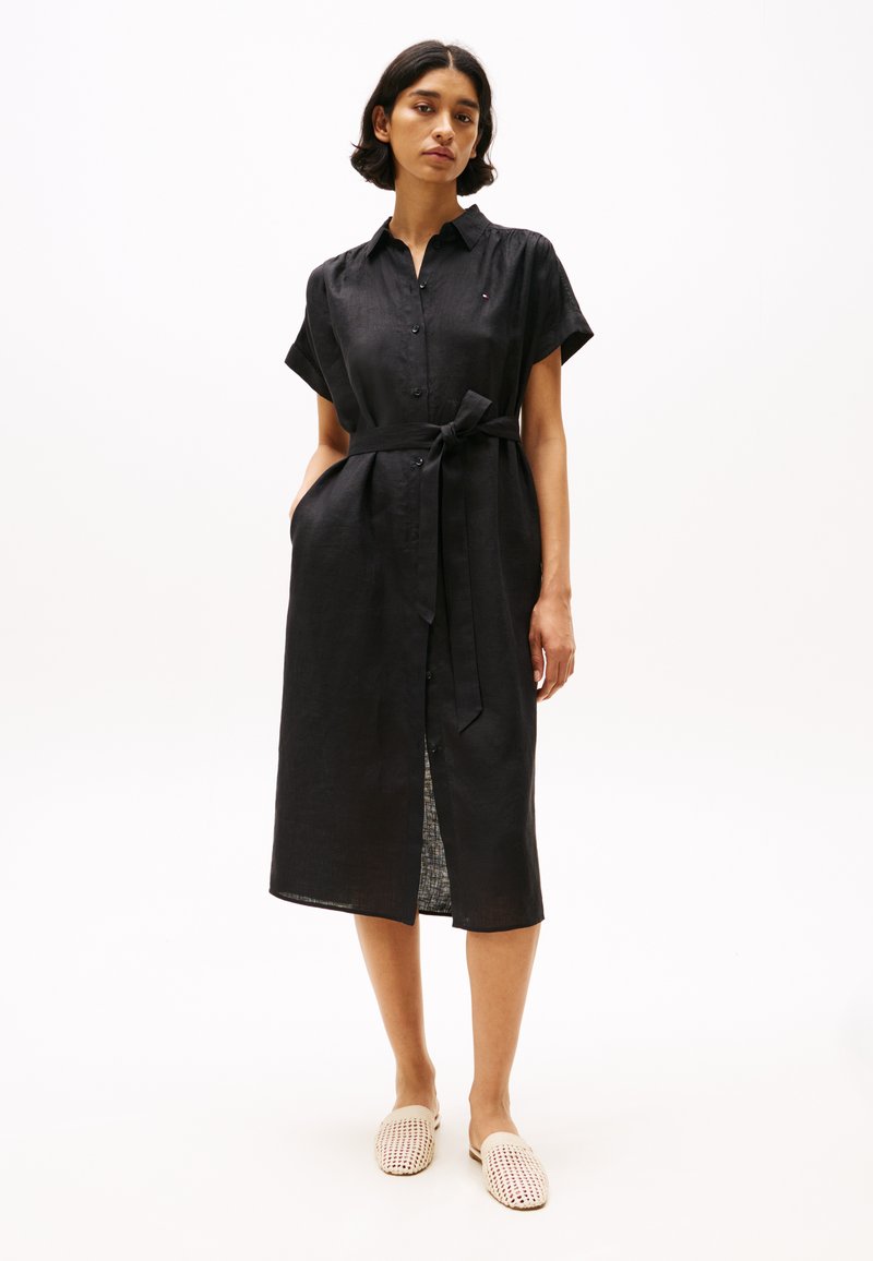 Rochie din in negru, cu mâneci scurte, închidere cu nasturi, talie cu curea, lungime până la genunchi și o fante. Asortată cu pantofi slip-on din material țesut ușor.
