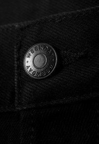 Svart denimstoff med en nærbildevisning av en sirkulær svart metallknapp gravert med "WEEKDAY." Teksturert overflate og synlige sømdetaljer.