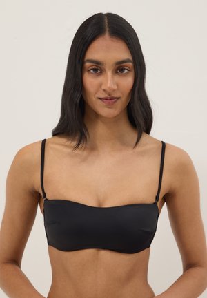 Frau mit langen schwarzen Haaren, die einen schwarzen Bandeau-BH mit dünnen verstellbaren Trägern trägt, posiert vor einem einfarbigen hellen Hintergrund.