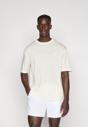 T-shirt beige chiaro a maniche corte con scollo rotondo, realizzata in tessuto morbido. Indossata con pantaloncini bianchi, presenta un dettaglio di logo discreto sul petto.