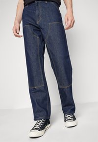 Dark blue denim pants with a relaxed fit, featuring distinct front stitching and pockets, paired with black sneakers and white laces.