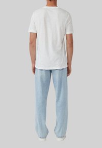 Eine Person trägt ein einfaches weißes Kurzarmhemd und bequeme hellblaue Jeans, steht mit dem Rücken zur Kamera vor einem grauen Hintergrund.