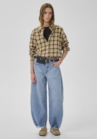 Skjorte i tern i gult og brunt, sort t-shirt, oversize lyseblå jeans med frynser nederst og brune slip-on sko. Sort bæltetaske ved taljen.