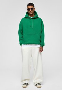Groene oversized hoodie met een voorzak, capuchon met trekkoord, gedragen over een wit shirt, gecombineerd met losse witte broeken en zwarte schoenen.