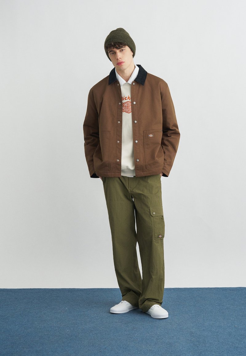 Young man wearing a brown jacket, white graphic hoodie, olive green cargo pants, olive beanie, and white sneakers standing on blue carpet.