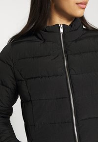 Giubbotto imbottito nero con colletto alto, chiusura frontale con zip e motivo trapuntato orizzontale. Il materiale ha una texture liscia e leggera.