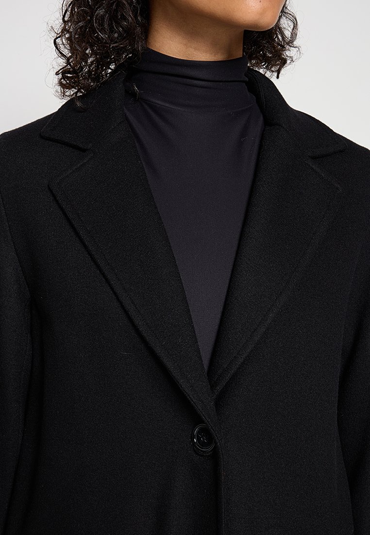 Schwarzer Wollblazer mit Reverskragen, einem Knopfverschluss und glatter Textur, über einem schwarzen Rollkragenoberteil getragen.