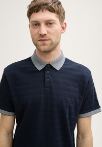 Granatowa koszulka polo z teksturowanymi paskami, jasnoszarym kołnierzykiem i zapięciem na trzy guziki. Krótkie rękawy z szarymi akcentami.