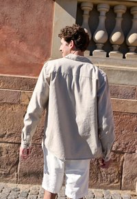 Svijetlosmeđi longsleeve majica s dugim rukavima i dugmadi na manžetama, opuštenog kroja, s detaljem na leđima, nosi se preko bijelih šortseva. Izgleda meko i izuzetno pamučno.