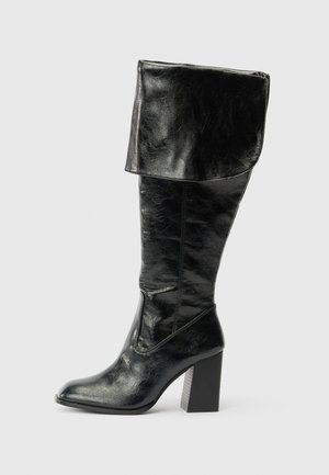 Bota de cuero negro hasta la mitad de la pantorrilla con la parte superior doblada y tacón cuadrado, mostrada de lado sobre un fondo blanco liso.