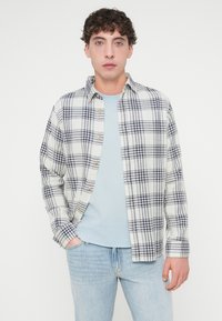 Καρό πουκάμισο σε εκρού και ναυτικό μπλε, βαμβακερό ύφασμα, σχεδιασμός με κουμπιά, φορεμένο πάνω από ανοιχτό μπλε μπλουζάκι, σε συνδυασμό με ανοιχτόχρωμα τζιν παντελόνια.