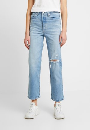 Frau trägt hellblaue, zerrissene Jeans mit geradem Bein und weiße Sneakers, steht vor einem einfarbigen weißen Hintergrund.