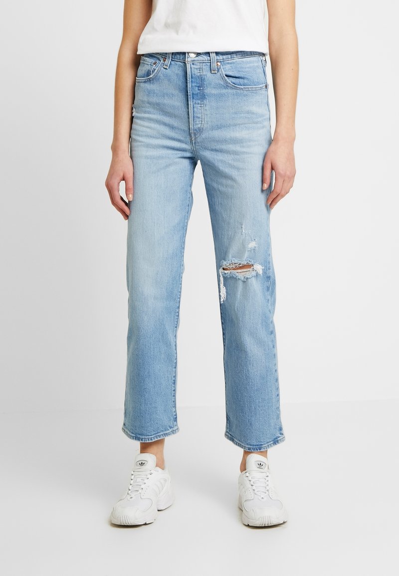 Femme portant un jean droit bleu clair déchiré et des baskets blanches, debout devant un fond blanc uni.