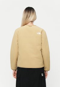 Veste rembourrée beige à manches longues, dotée d'un logo blanc dans le haut du dos, conçue avec un col rond et une texture lisse.