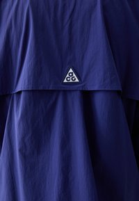 Marineblå yttertøy med en brettet krage, dekorert med en trekantet logo-patch i hvitt. Glatt tekstur, lett stoff.